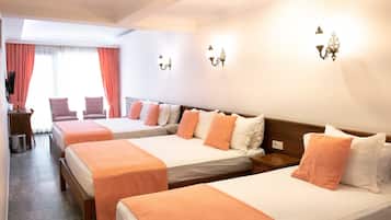 Habitación de hotel con cuatro camas individuales, cada una con cubrecama blanco y naranja, y cabecero de madera. Hay dos lámparas de pared y una ventana con cortinas.