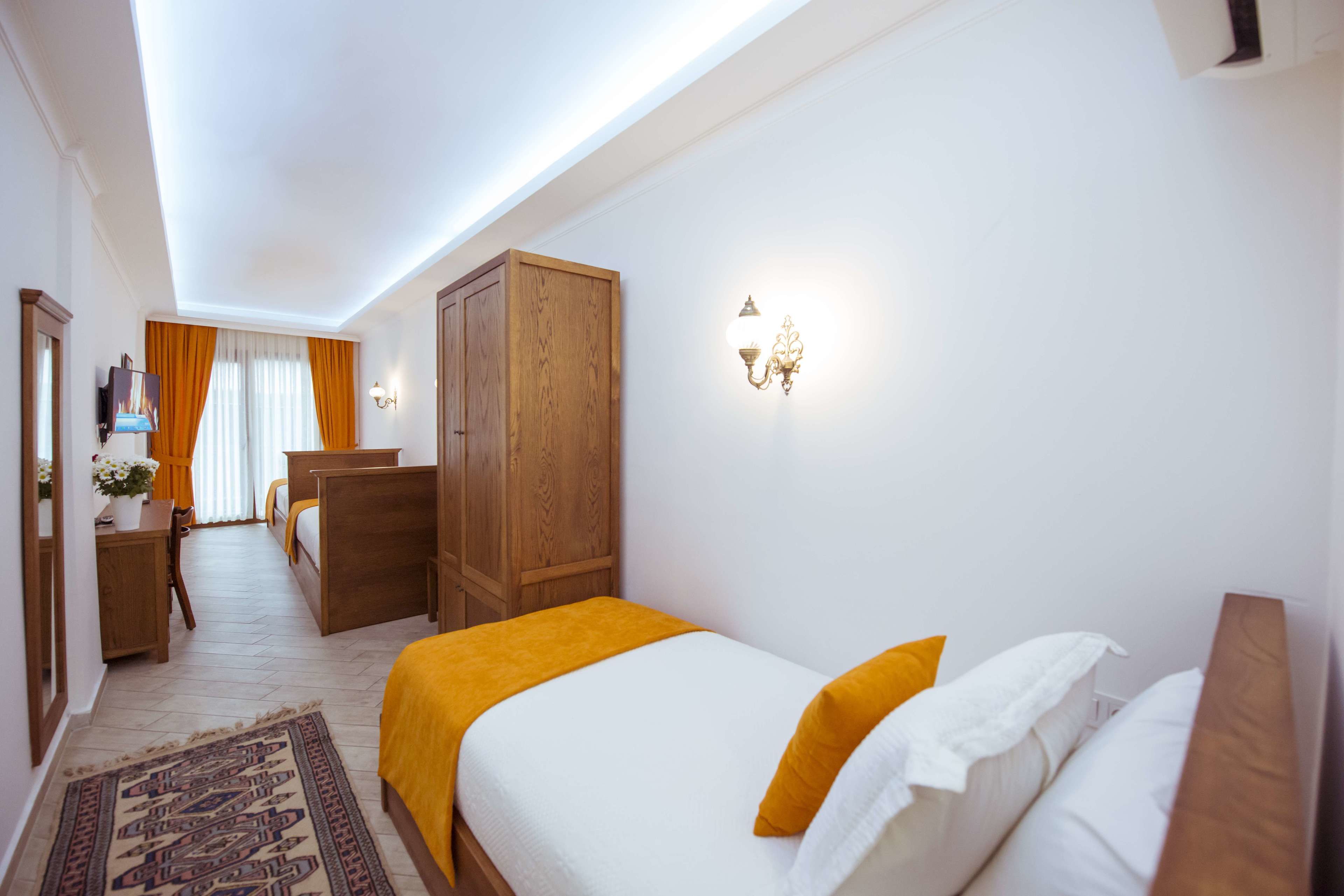 A hotel room with a bed, a wooden wardrobe, a desk with a chair, a small table with a plant, and a large window with curtains.