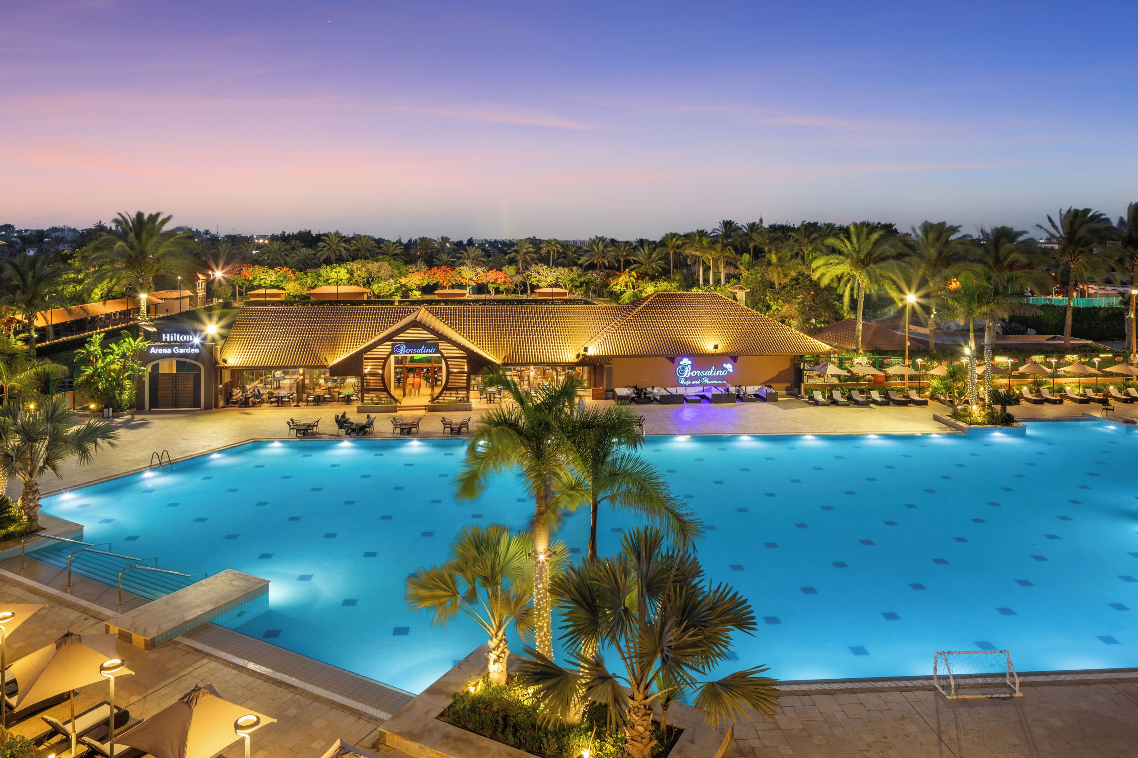 Una gran piscina rodeada de palmeras y sillas de descanso, con un edificio identificado como 'Hilton Arena Garden' al fondo.