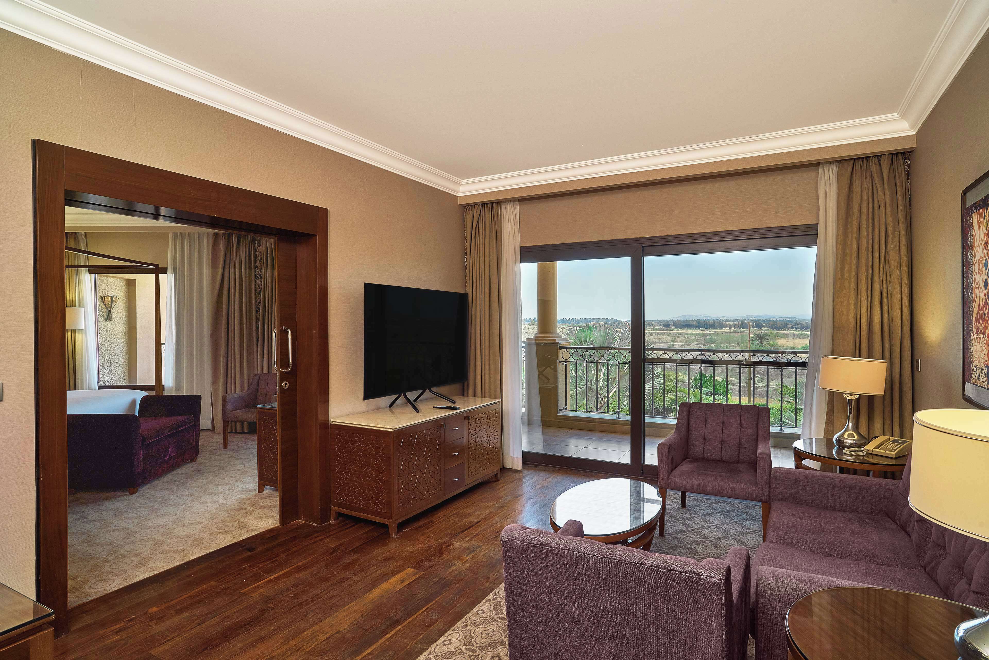 A hotel room with a large balcony, a flat-screen TV, a sofa, a coffee table, and a desk with a lamp.