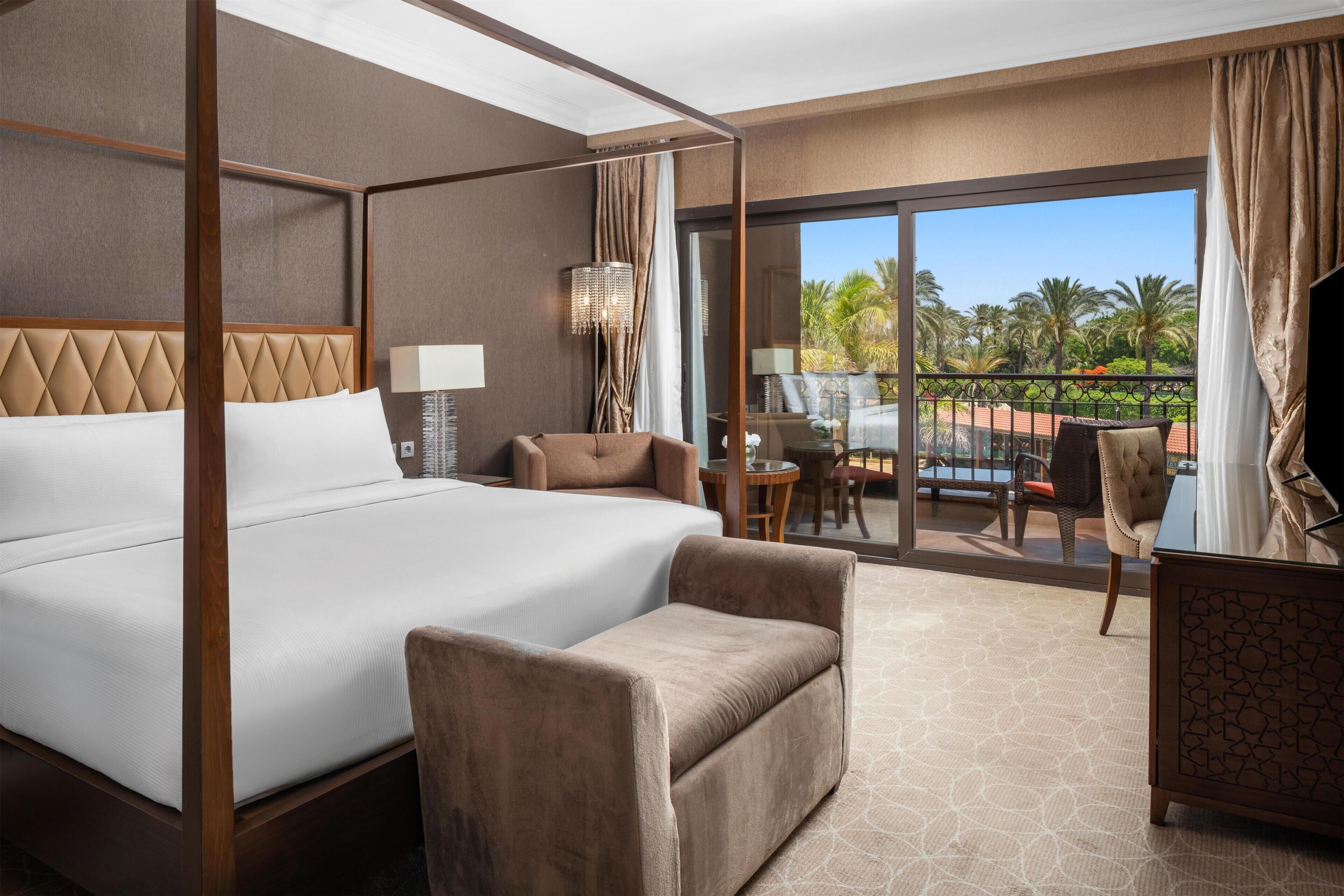 A hotel room with a large bed, a sitting area with a chair and a table, a TV, and a balcony with a view of palm trees.