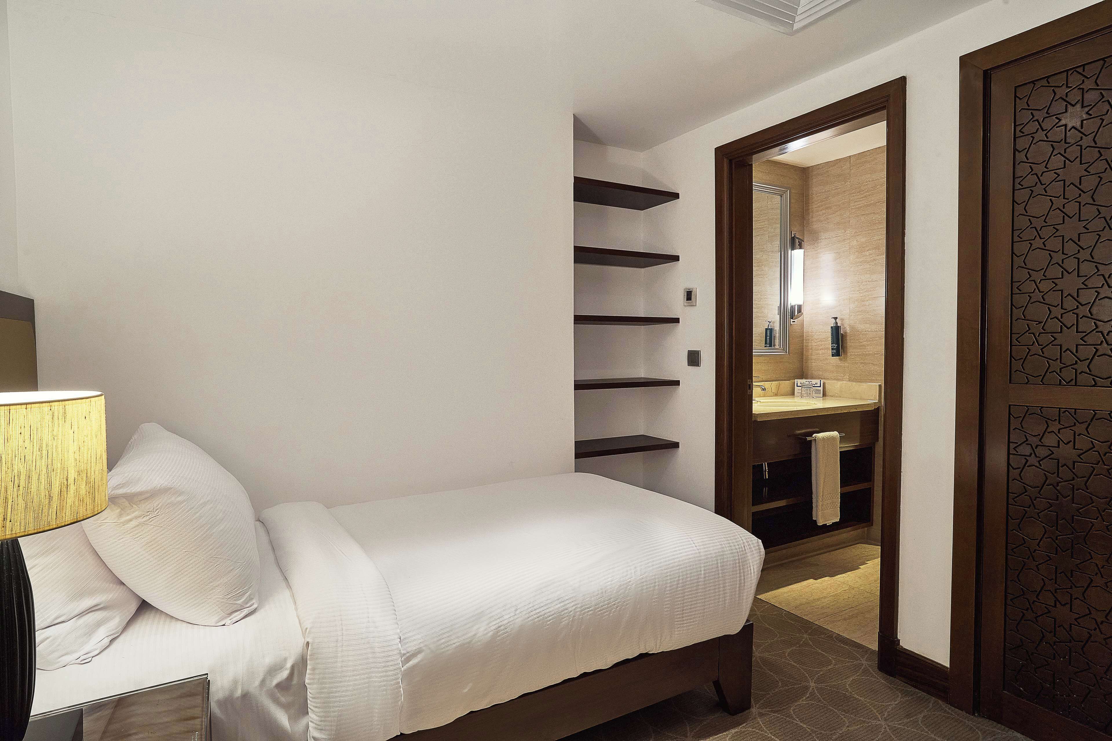 A bedroom with a bed, a bedside lamp, a mirror, and a bathroom visible through an open door.