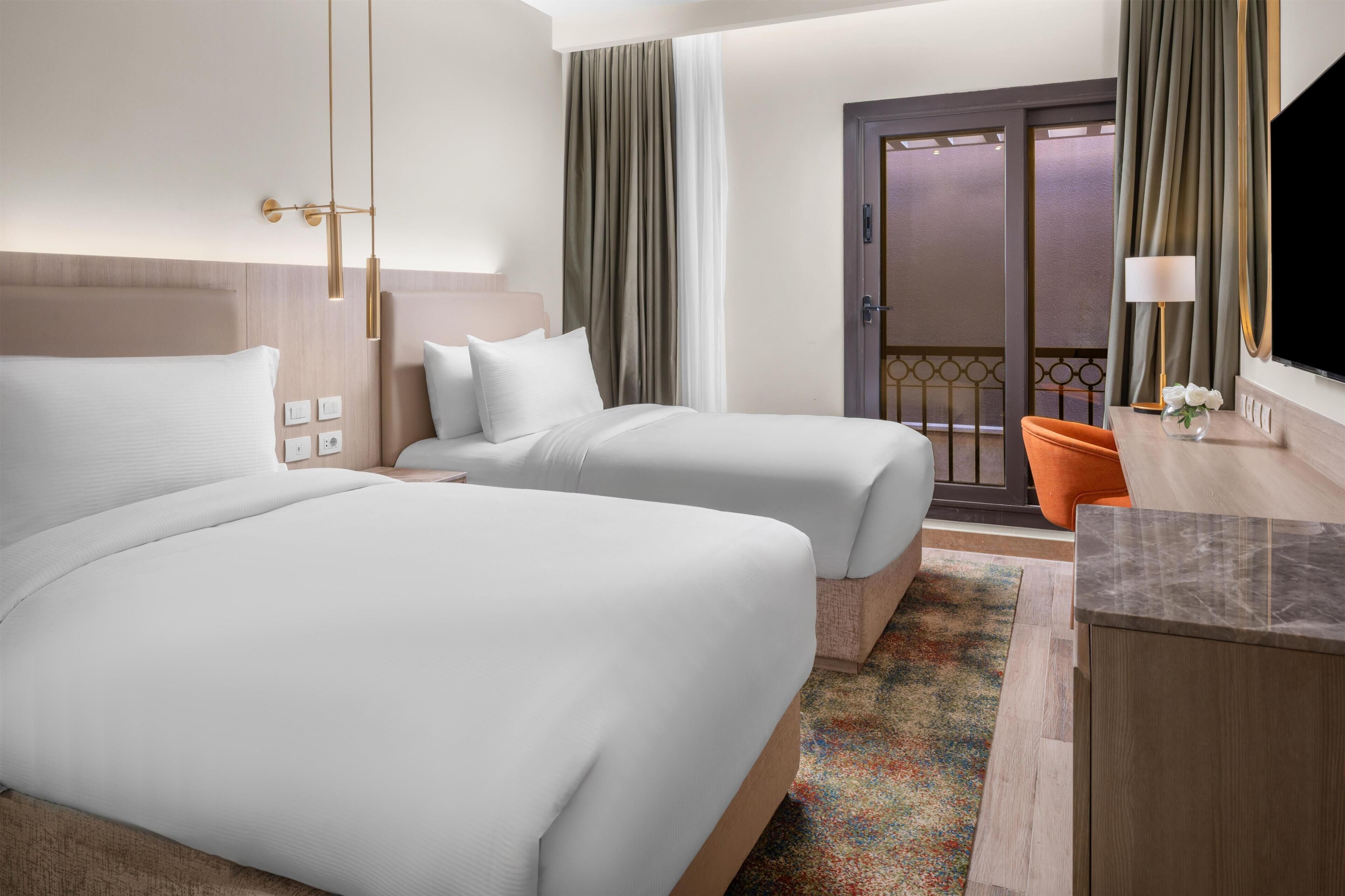 A hotel room with two beds, a desk, a chair, a TV, and a balcony with curtains.