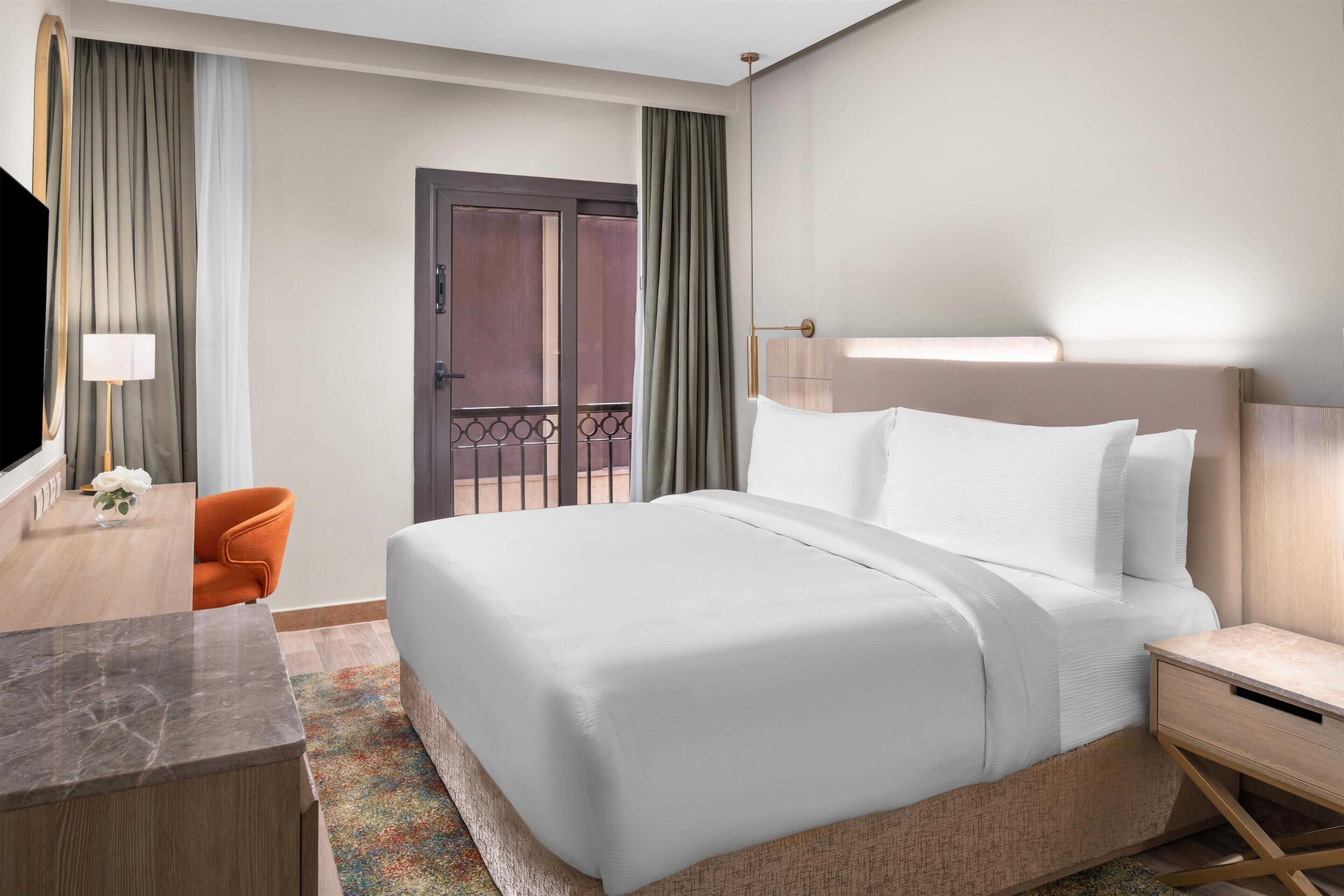 A hotel room with a bed, a desk, a chair, a TV, and a door leading to a balcony.