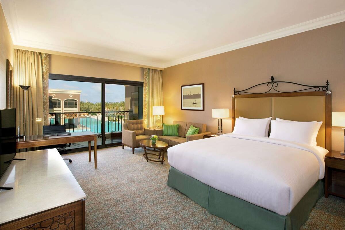 A hotel room with a bed, a desk, a chair, a small table, and a view of a pool.