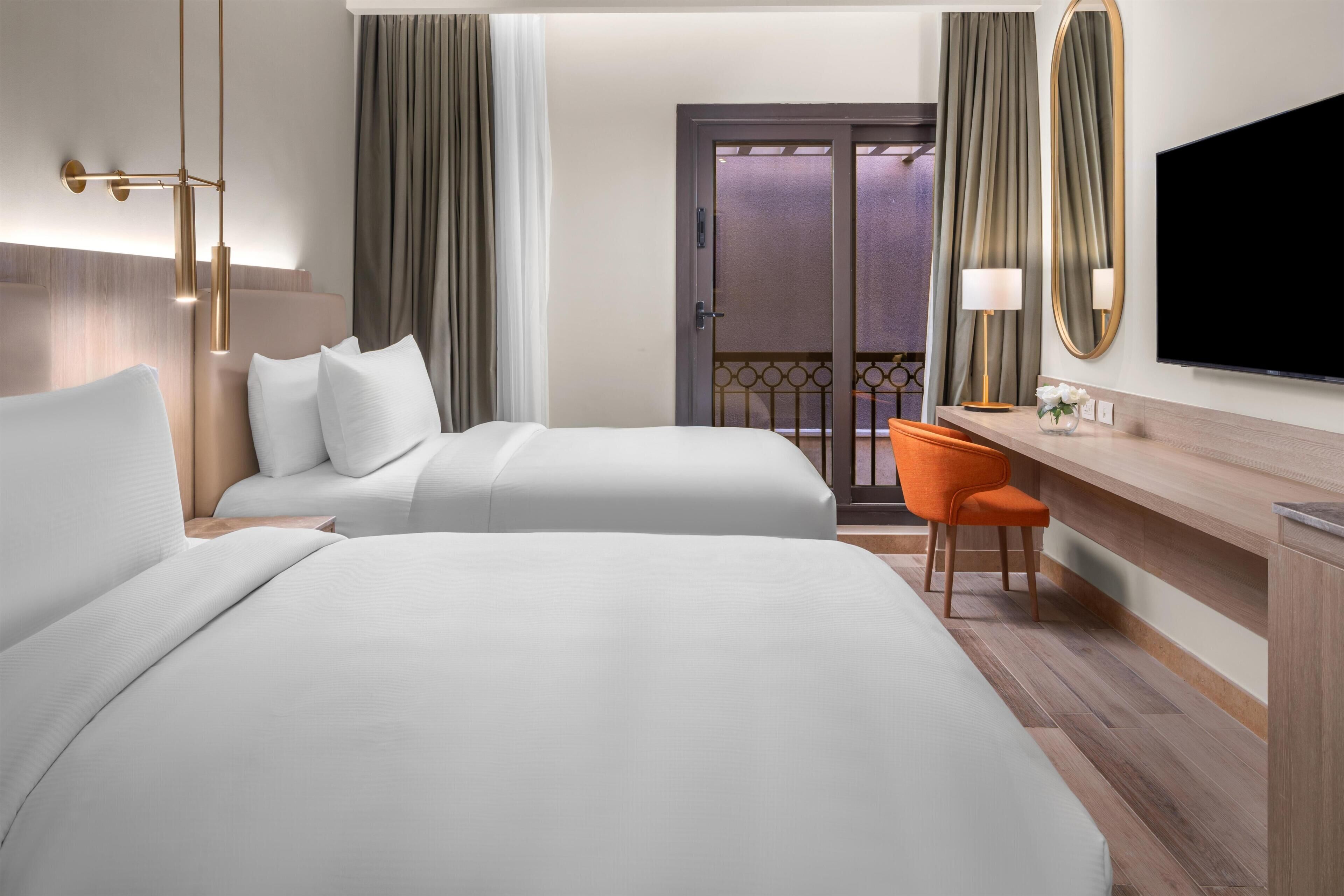 A hotel room with two beds, a desk, a chair, a television, and a balcony with curtains.