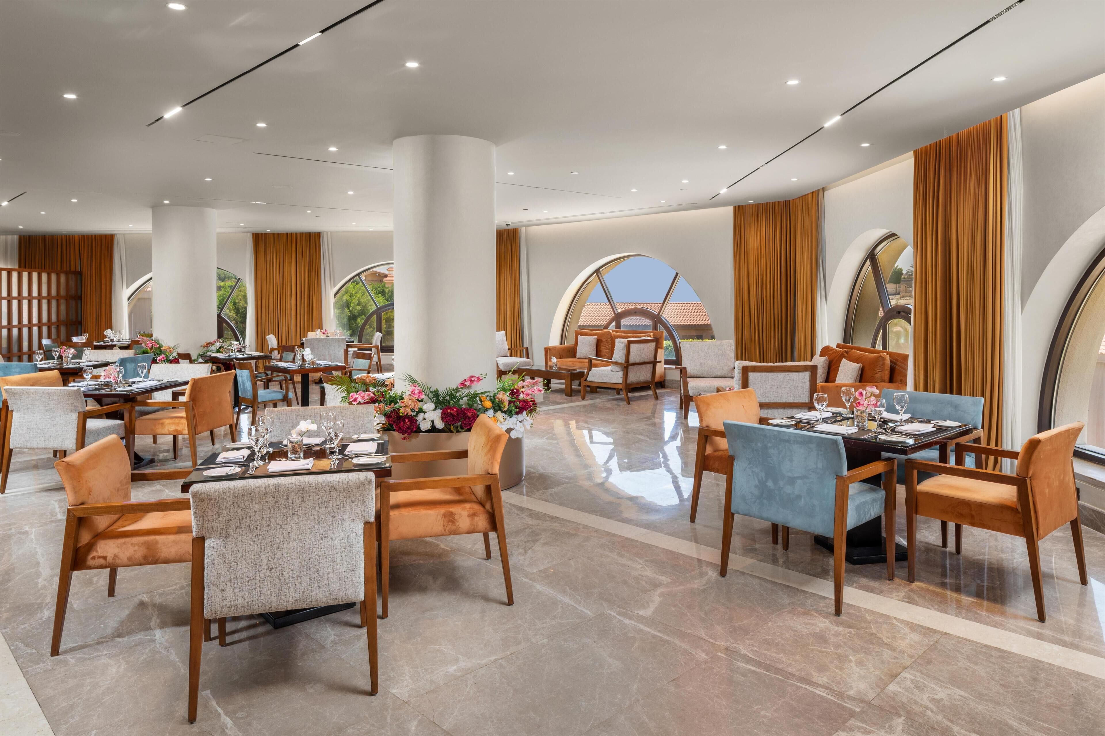 A spacious hotel dining area with multiple tables and chairs, large windows, and a modern interior design.