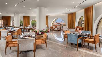 A spacious hotel dining area with multiple tables and chairs, large windows, and a modern interior design.