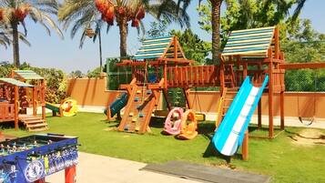 Explore the outdoor play area, featuring a multi-level playhouse, slides, and swings, surrounded by a fence and a foosball table.