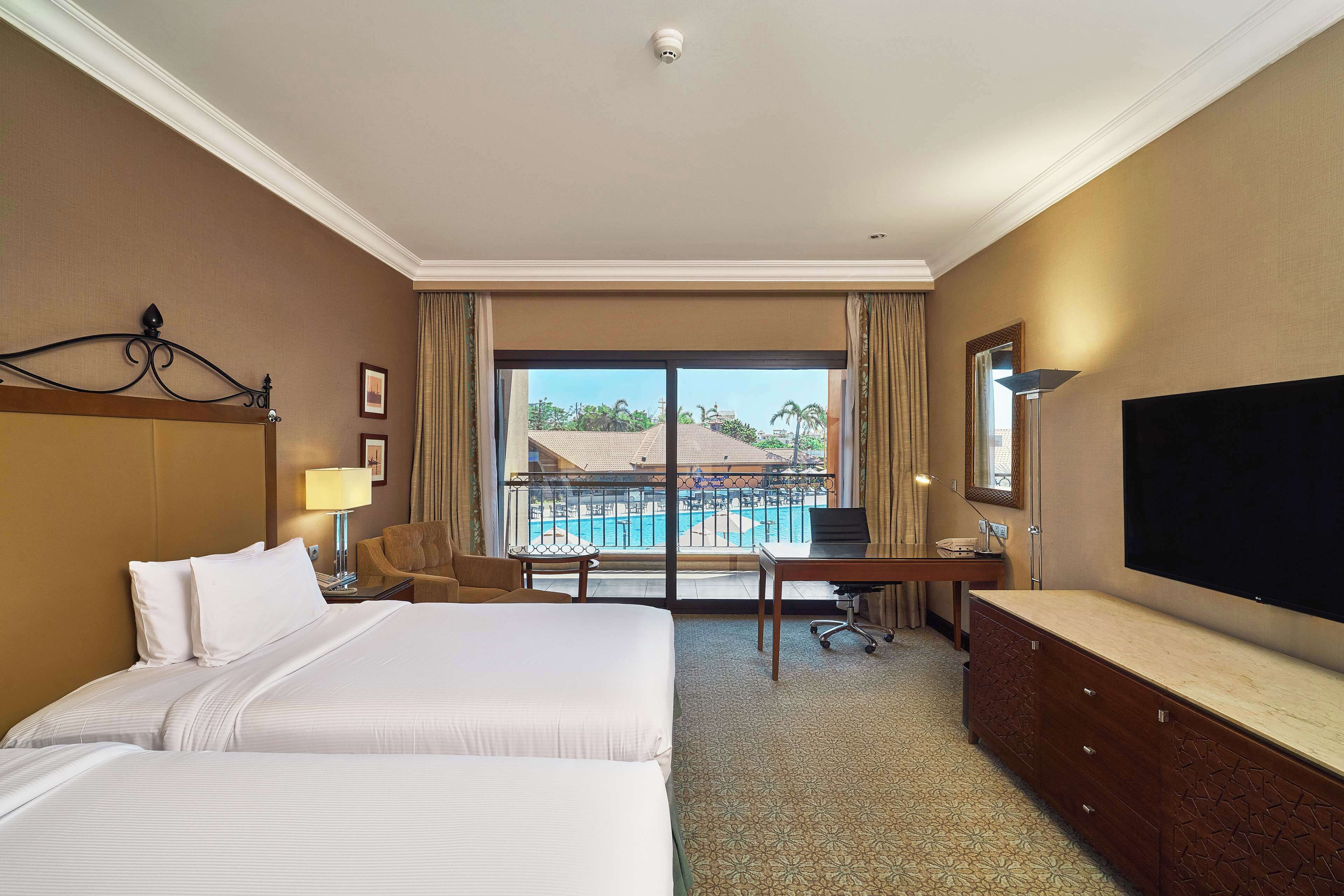 A hotel room with two beds, a desk, a TV, and a view of a pool.