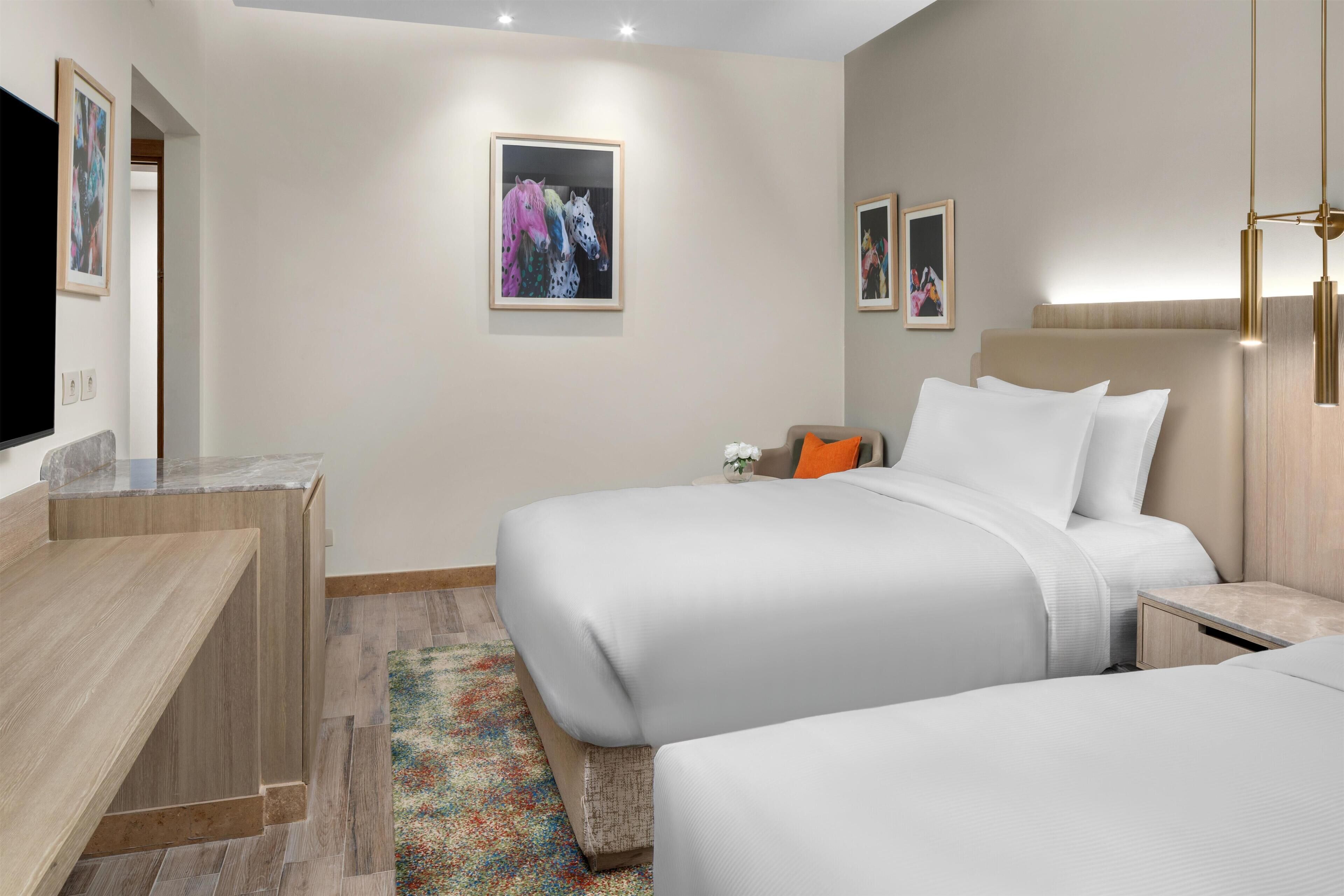 A hotel room with two beds, a desk, a television, and artwork on the walls.