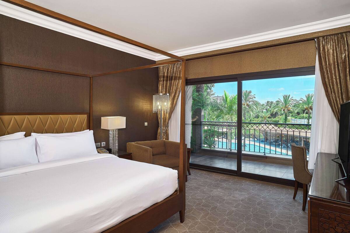 Une chambre d’hôtel dotée d’un grand lit, d’un coin salon, d’un balcon avec vue sur la piscine et d’une télévision.