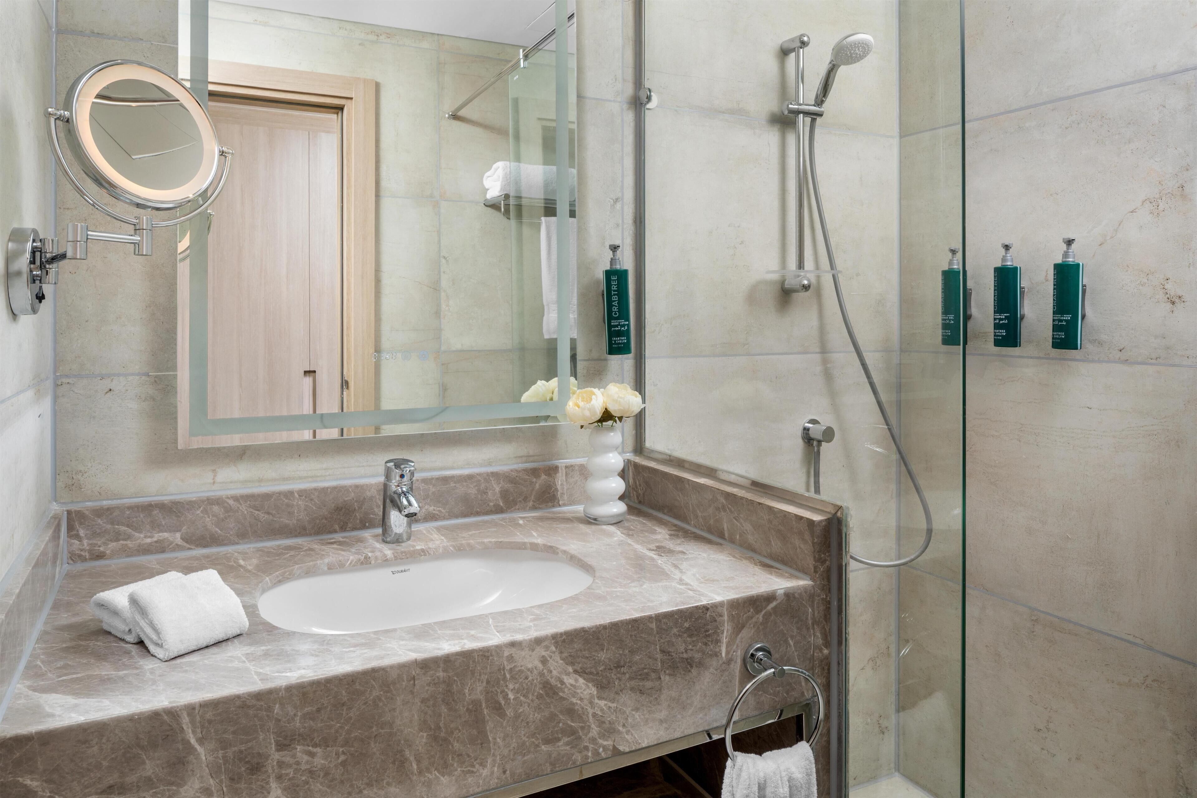 A bathroom with a marble sink, a round mirror, a glass shower enclosure, and toiletries on the wall.