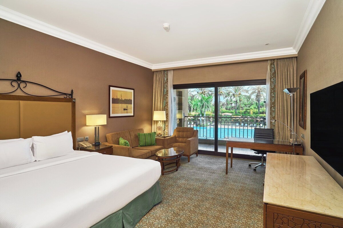A hotel room with a bed, a desk, a chair, a sofa, a TV, a lamp, and a balcony with a pool view.