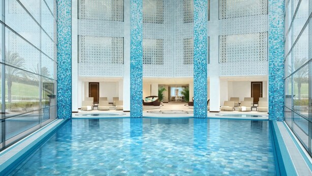 An indoor swimming pool with clear blue water, surrounded by glass walls and decorative blue-tiled pillars.