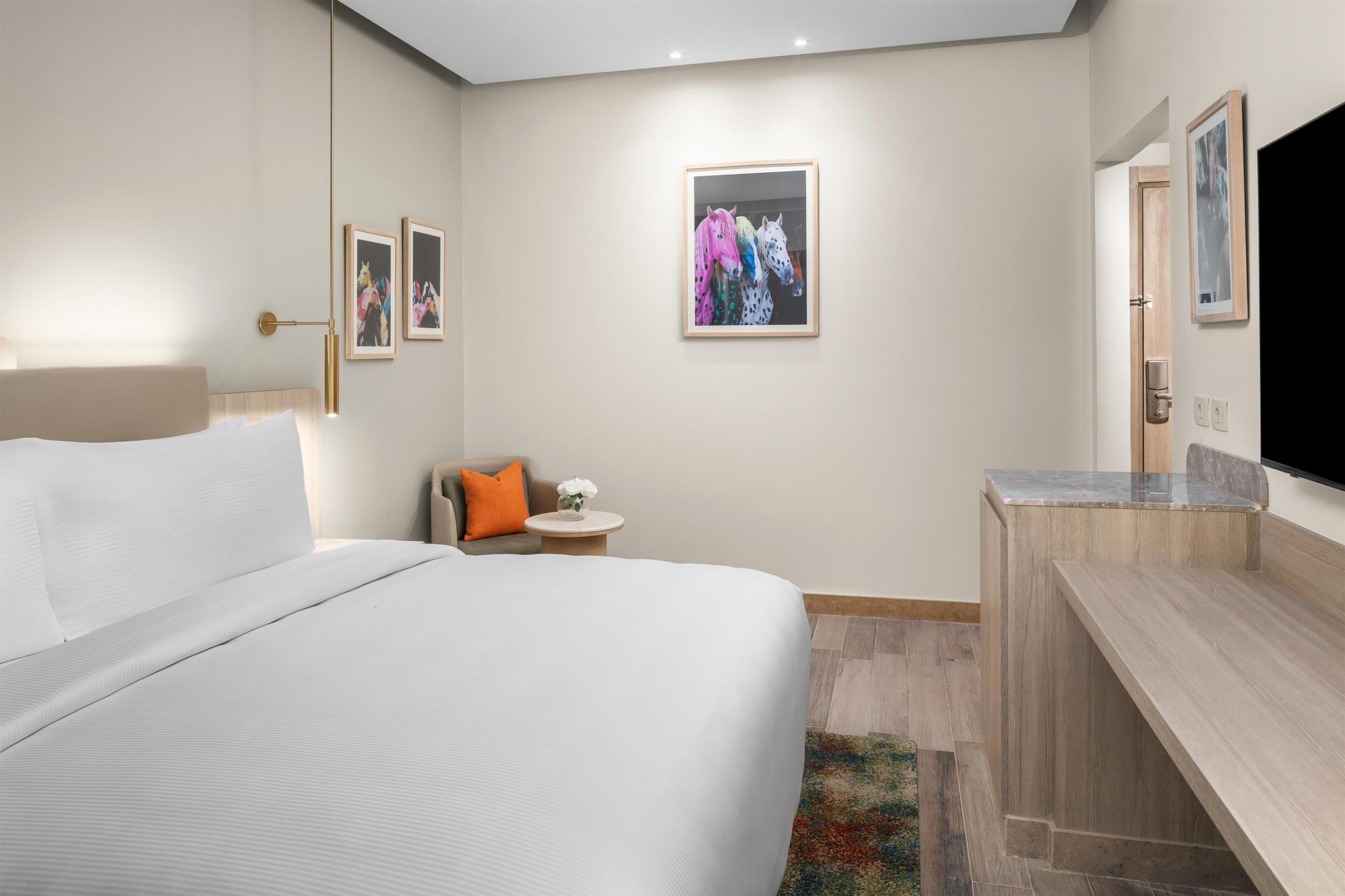 Une chambre d’hôtel avec un lit, une petite table ronde ornée d’un vase de fleurs, un canapé avec un coussin orange et un téléviseur fixé au mur.