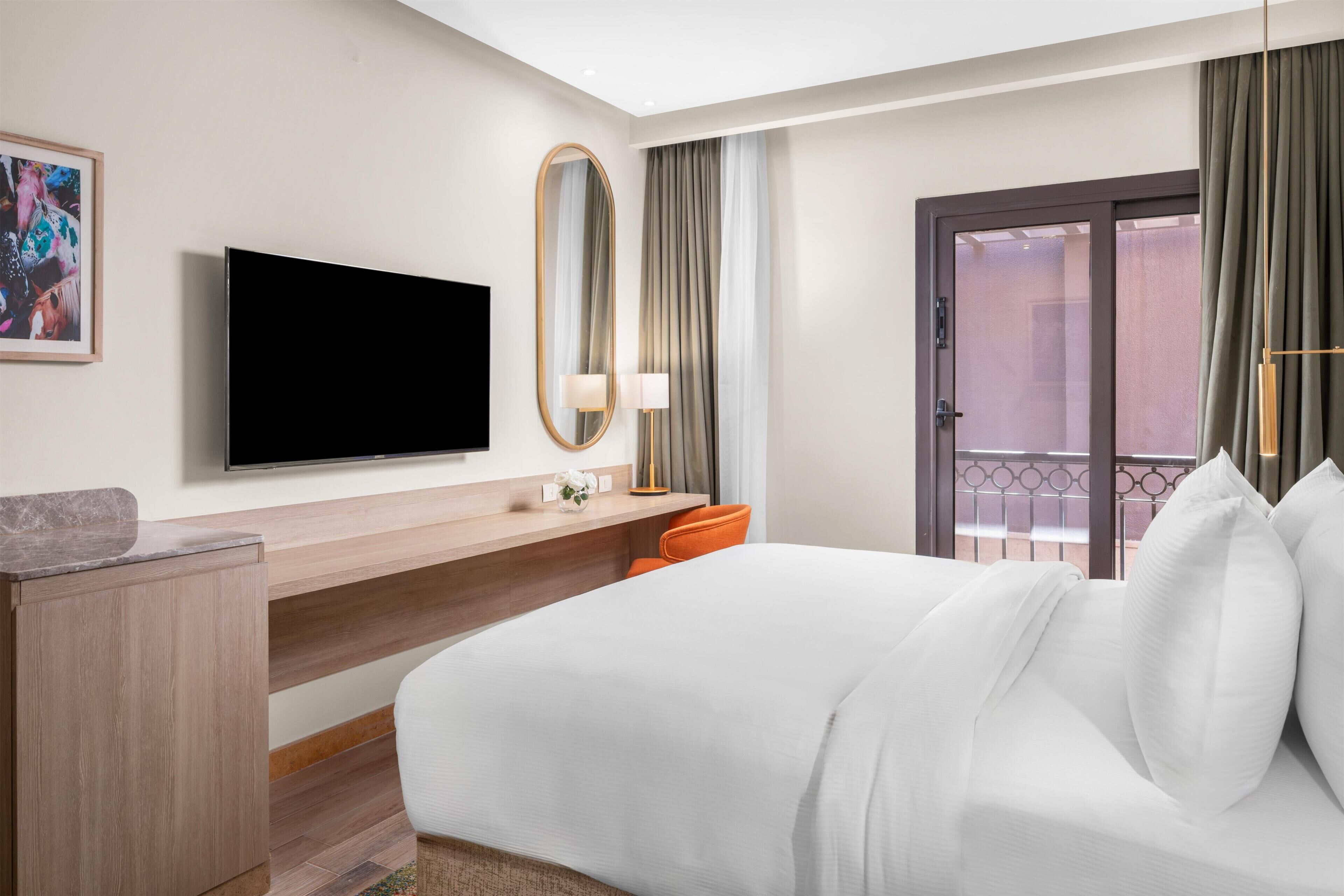 Een hotelkamer met een bed, een aan de muur bevestigde televisie, een bureau met lamp en een raam met gordijnen.