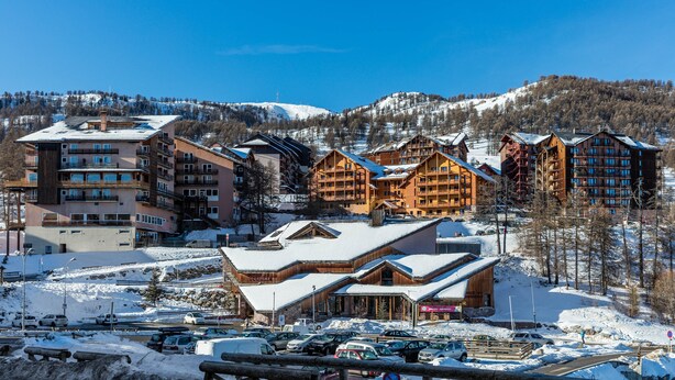Une station de ski dotée de plusieurs chalets en bois, d’un parking et offrant une vue imprenable sur des montagnes enneigées.
