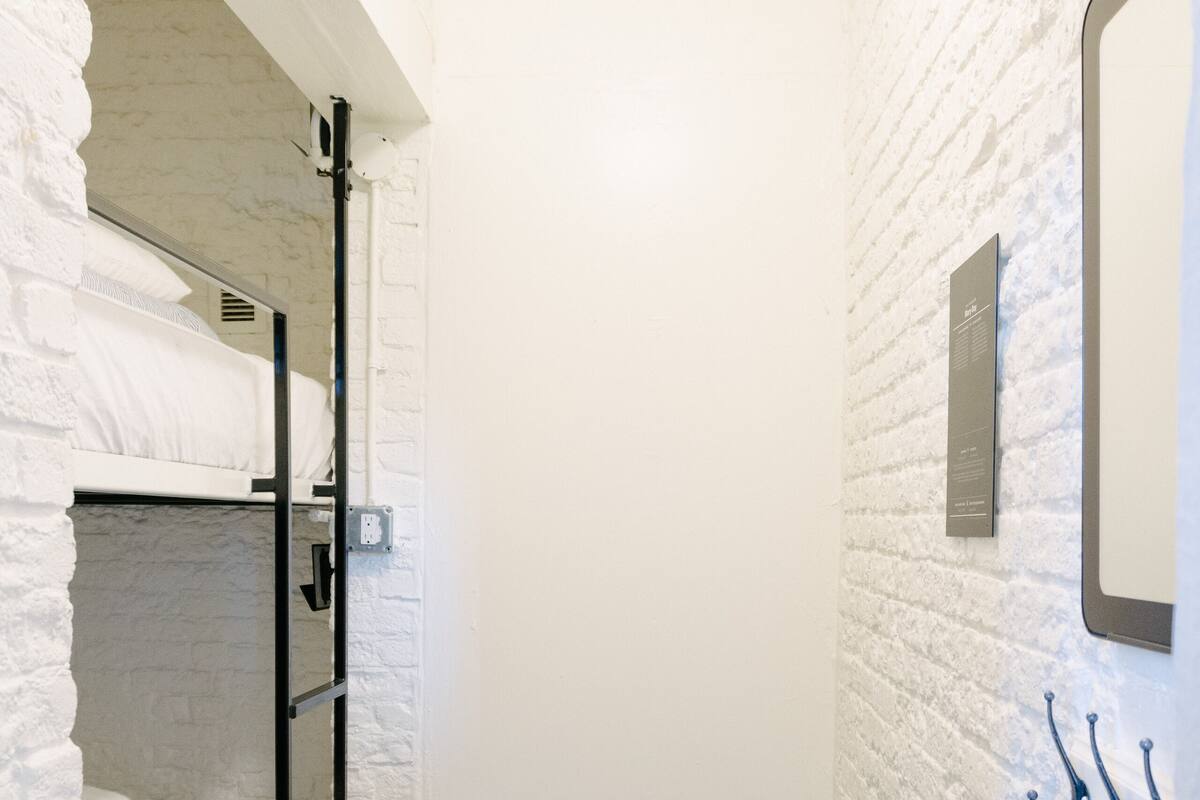 A narrow room with white brick walls, a bunk bed, a small stool, and a mirror on the wall.