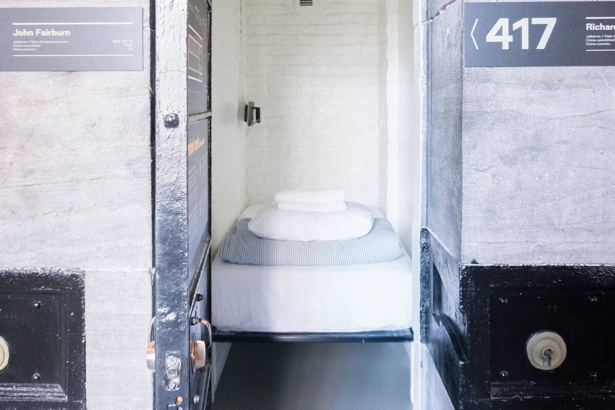 A narrow, enclosed passageway leading to a room with a bed, a small window, and a door with a number plate.