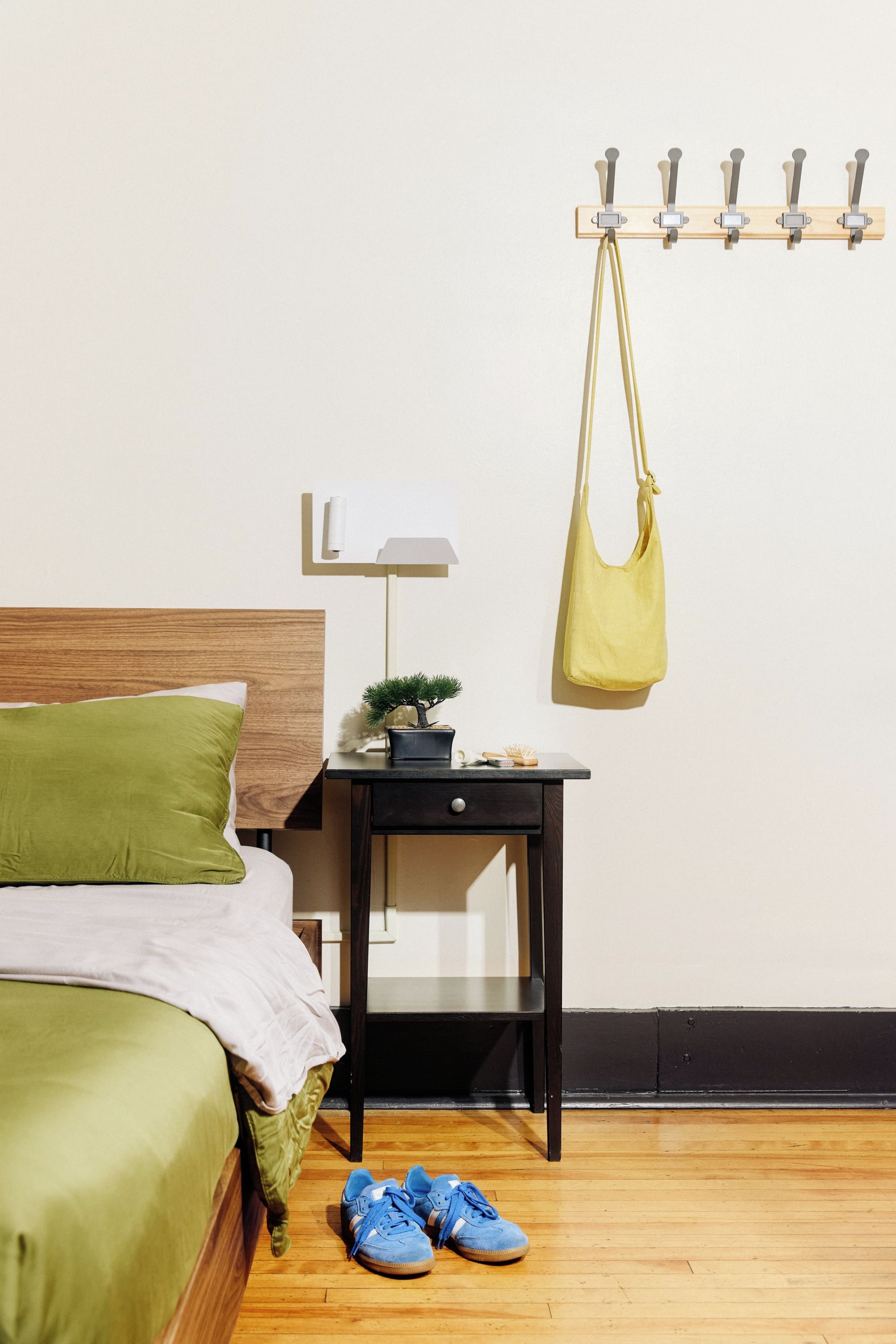 Un dormitorio con cama, mesita de noche con lámpara, una planta pequeña y un gancho con un bolso.