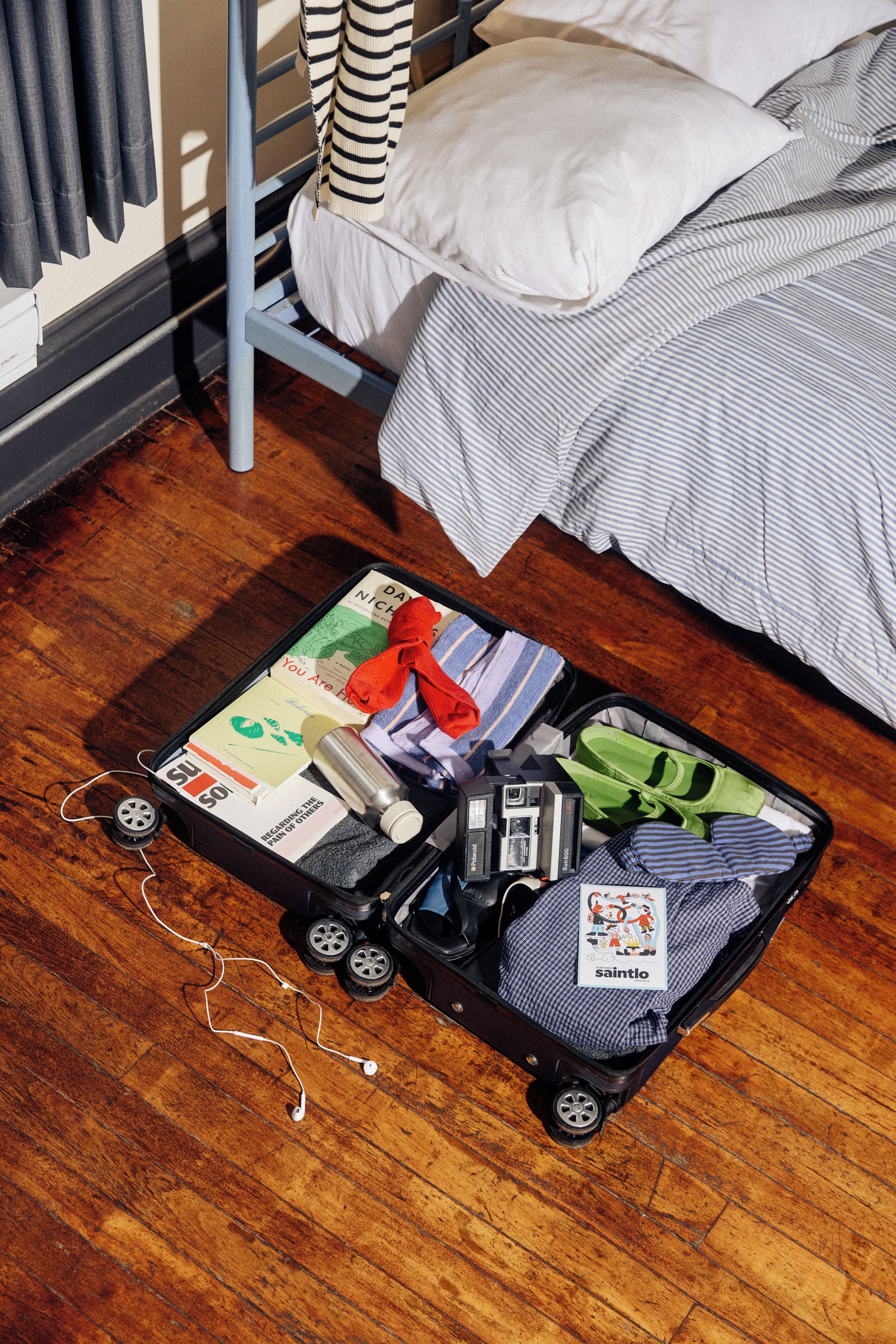 A suitcase on a wooden floor with various items including a book, a camera, and a pair of earphones.