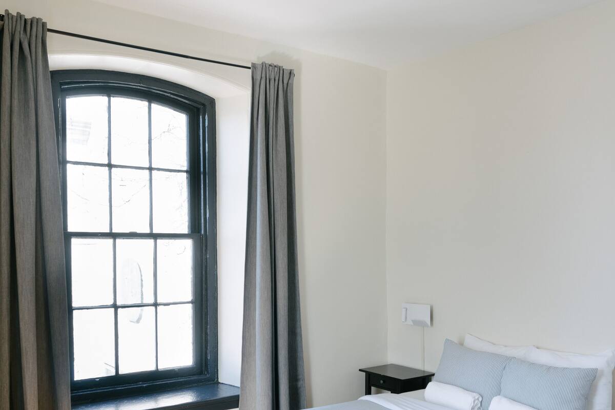 A bedroom with a bed, a nightstand, a window with curtains, and a thermostat on the wall.