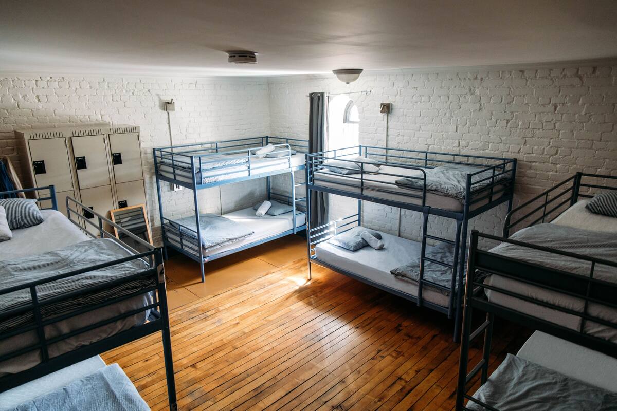 A dormitory room with bunk beds, wooden floors, white brick walls, and a window with curtains.
