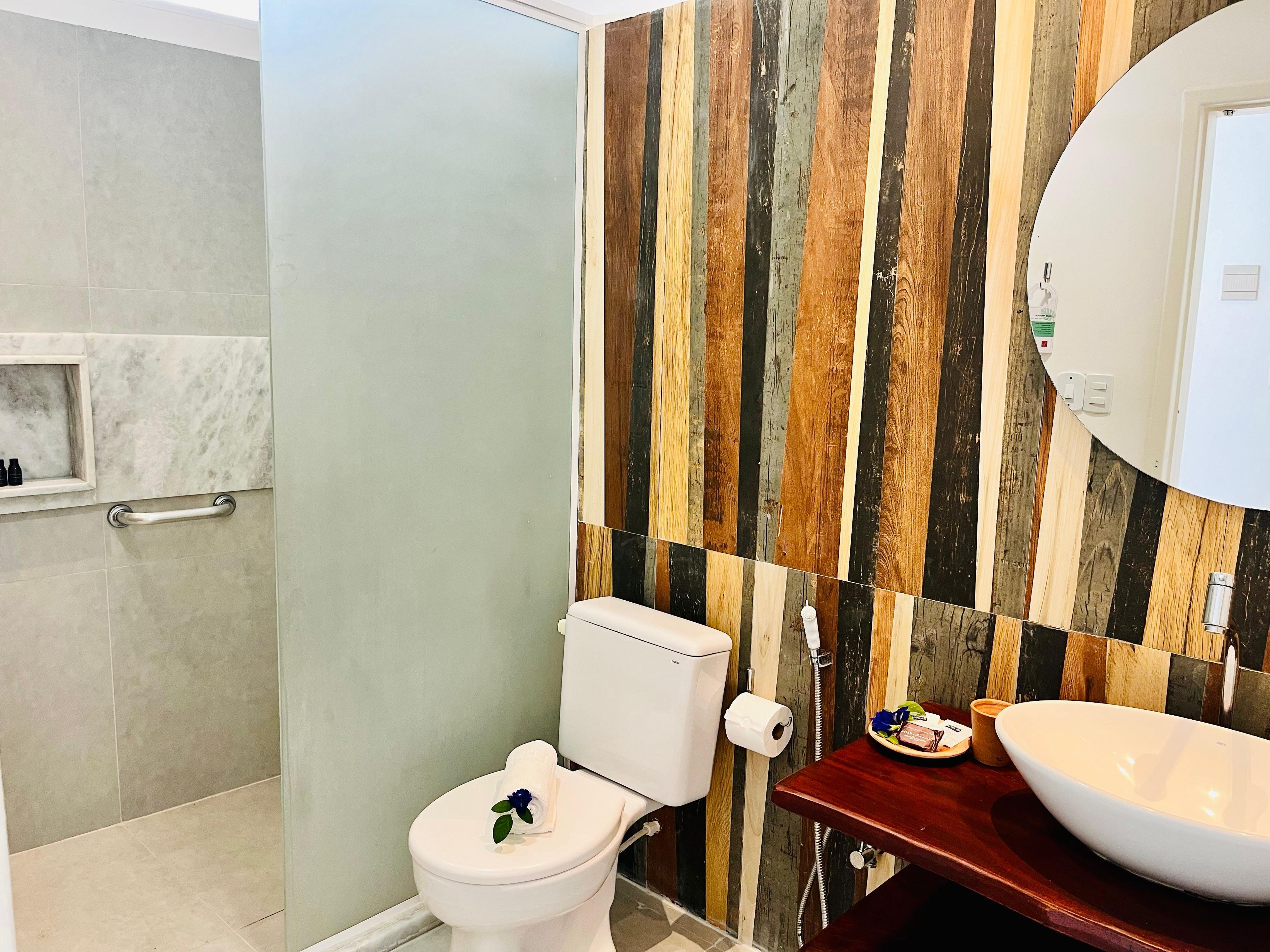 Un baño con inodoro blanco, pared de paneles de madera, zona de ducha con azulejos de mármol y tocador de madera con un lavamanos grande.