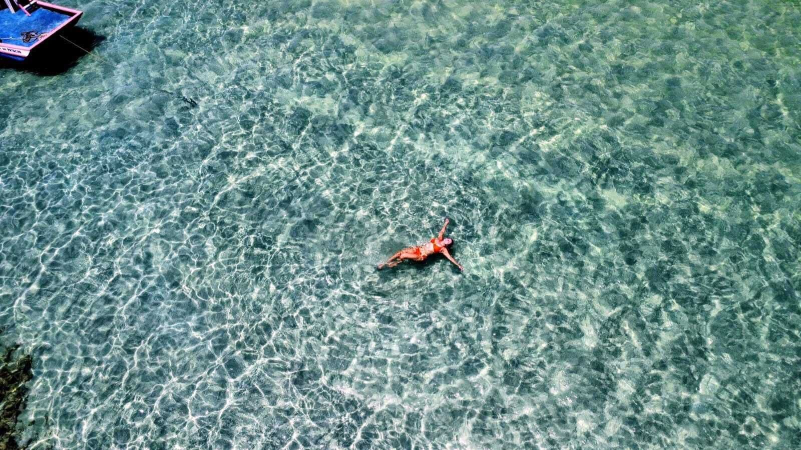 Una persona flota en agua cristalina y poco profunda, con un bote al fondo.