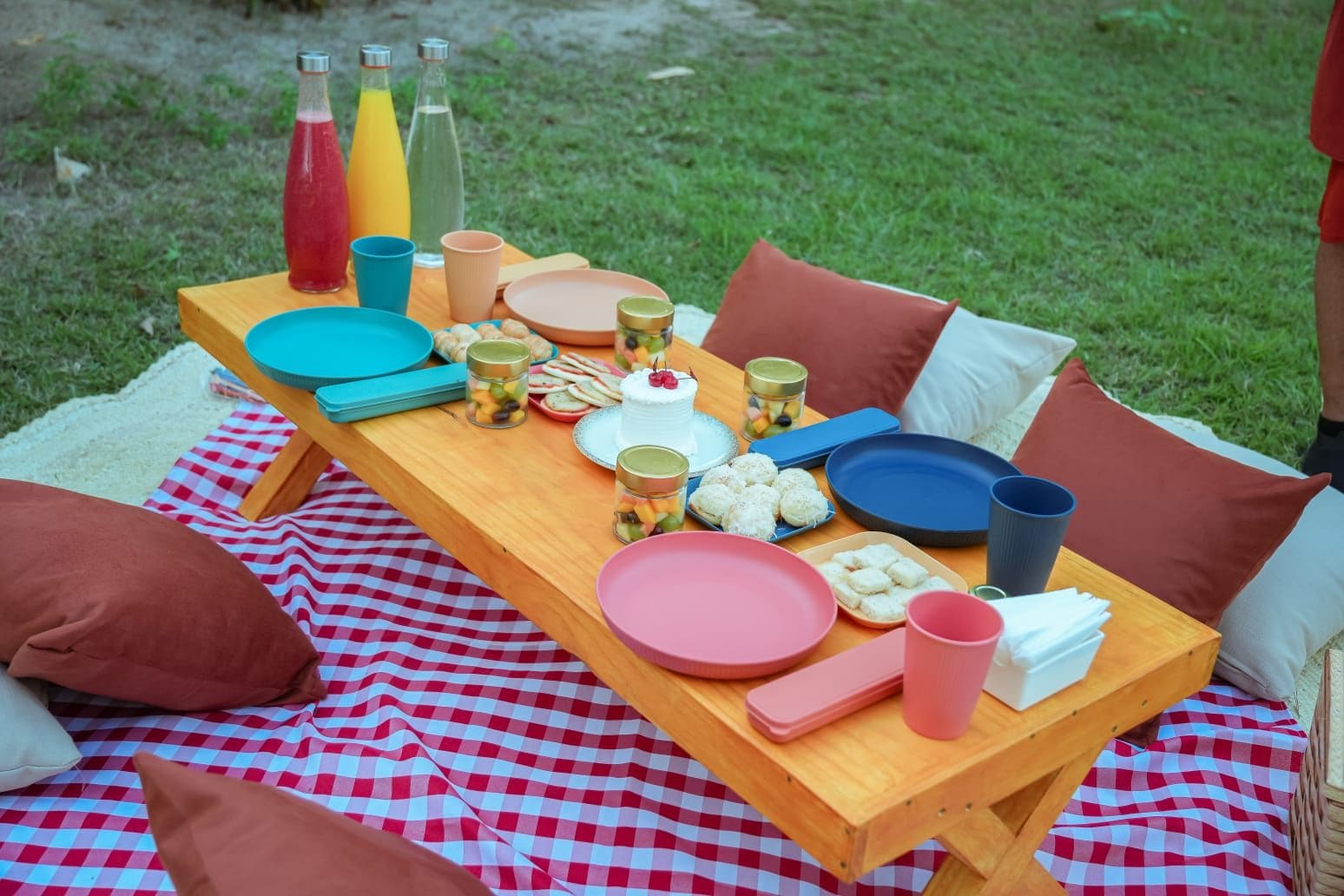 A picnic setup with a checkered blanket, a wooden table, and various food items and drinks.