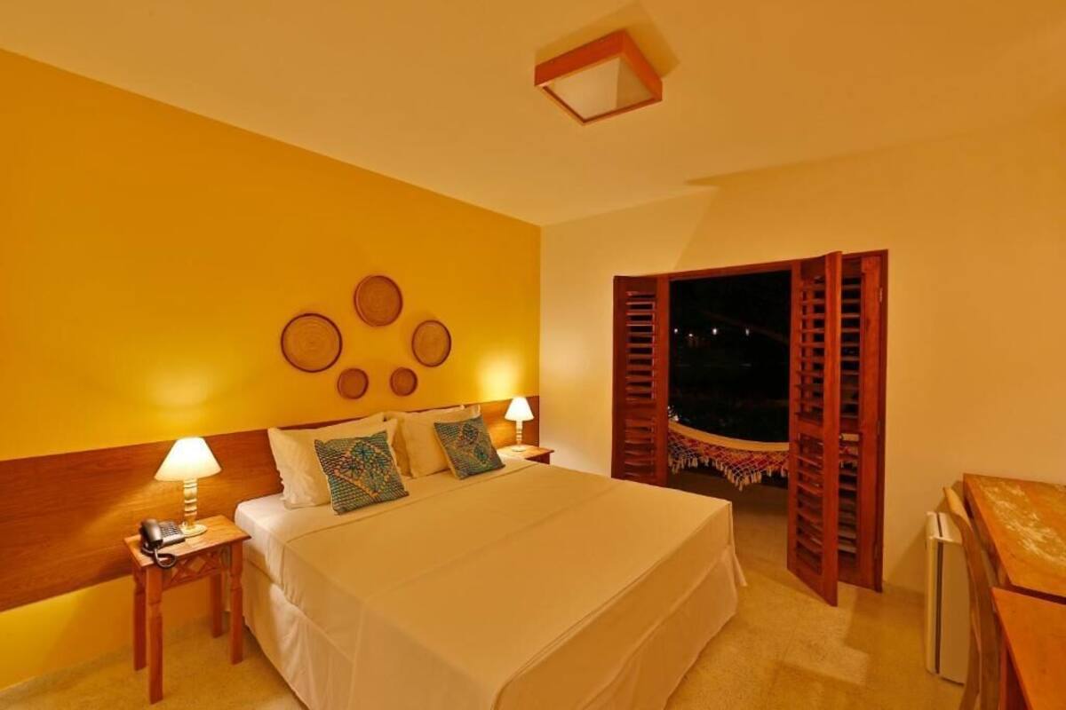 Hotelkamer met een groot bed, twee nachtkastjes met lampen, een bijzetmeubel en uitzicht door openslaande luiken.