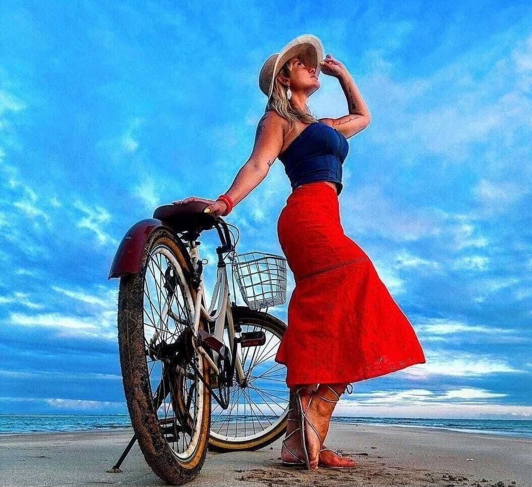 Una mujer con falda roja y blusa azul apoyada en una bicicleta en la playa, con el océano al fondo.