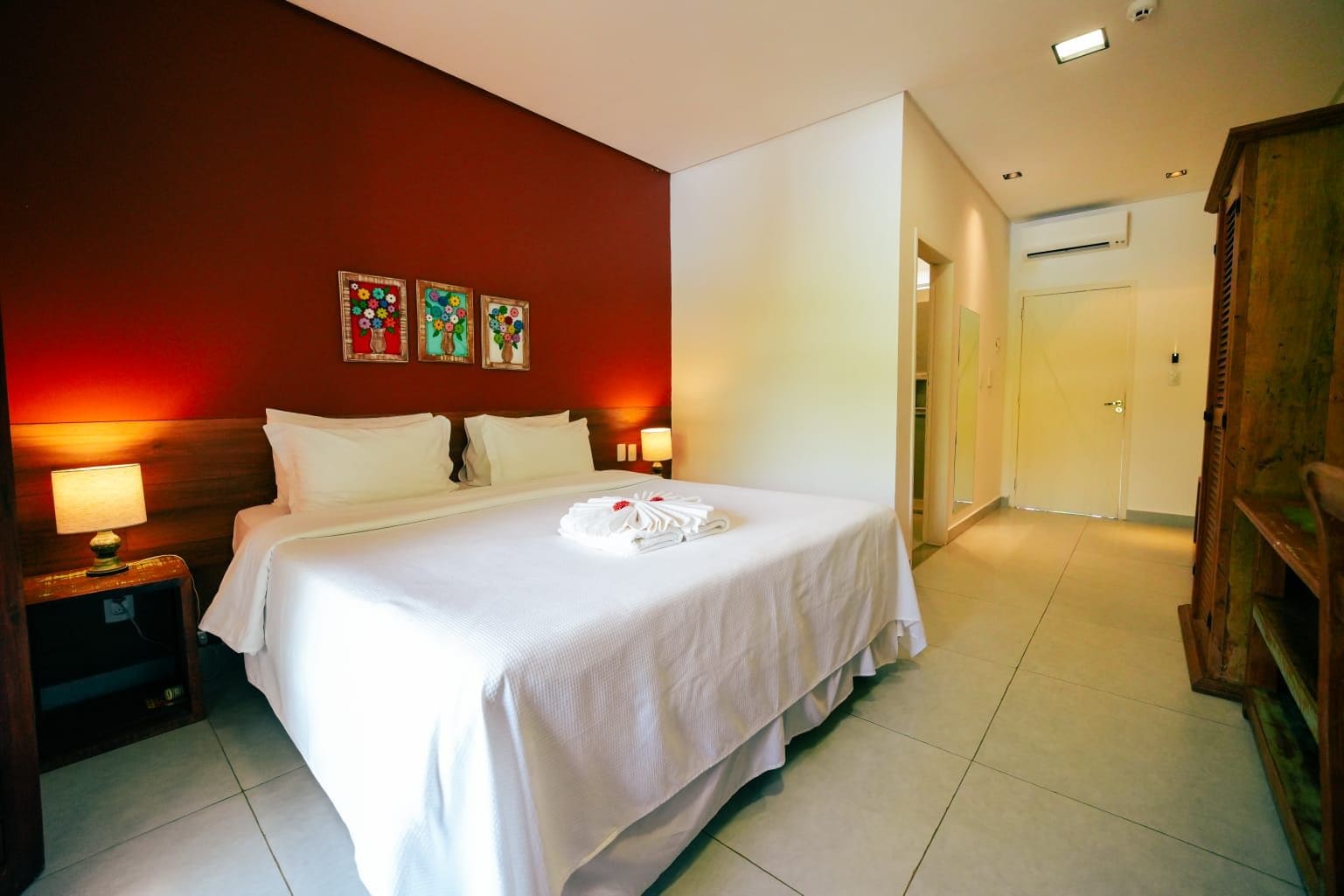 A hotel room with a large bed, two bedside tables with lamps, a wooden wardrobe, and a red accent wall with floral artwork.