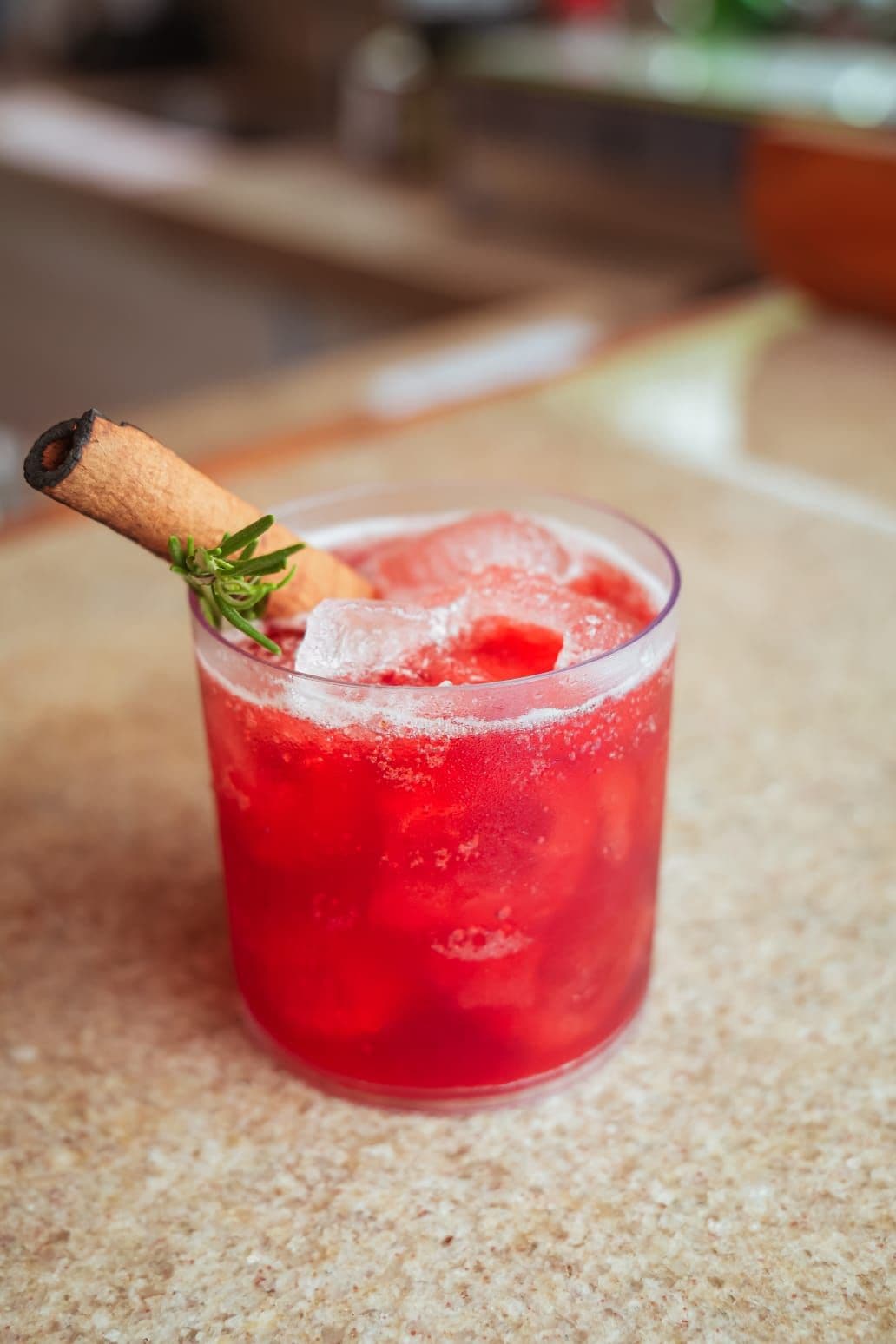 Un vaso con una bebida roja, hielo y una ramita de canela sobre una superficie texturizada.