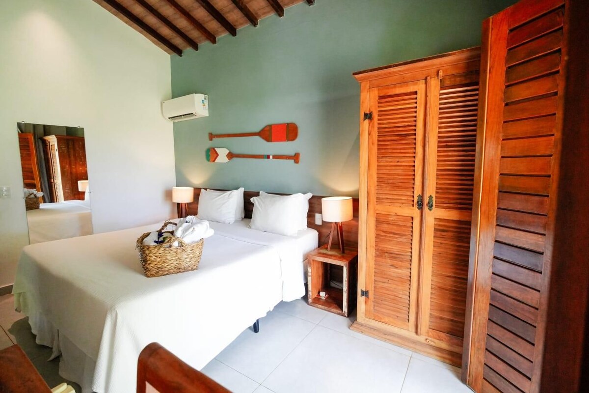 A hotel room with a bed, a wooden wardrobe, a wall-mounted air conditioner, and a small table with a lamp.