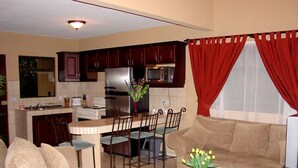 A living room with a sofa, a coffee table with a vase, and a kitchen area with a dining table and chairs. - Villas Roma (Santiago)