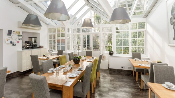 A bright dining area with a long wooden table set for six, surrounded by grey upholstered chairs. Large windows allow natural light to fill the space, and a skylight above enhances the brightness. - 94DR (Edinburgh)