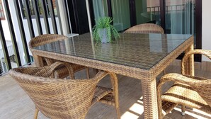 Une table à manger avec un plateau en verre, des chaises en osier, une plante en pot et une vue sur un bâtiment à travers une porte vitrée.