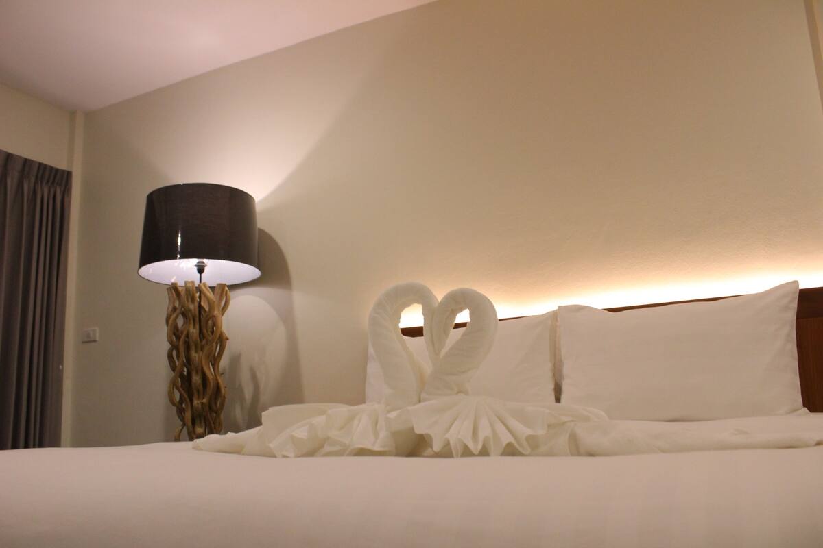 Un lit bien fait, avec du linge de lit blanc et un arrangement décoratif en forme de cygne. Une lampe moderne, dotée d’un pied en bois original, est placée sur le côté gauche.