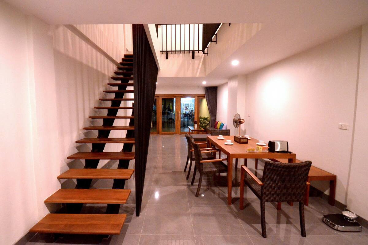 Un escalier menant à un deuxième étage, un coin repas avec des meubles en bois et une cuisine visible à travers une porte ouverte.