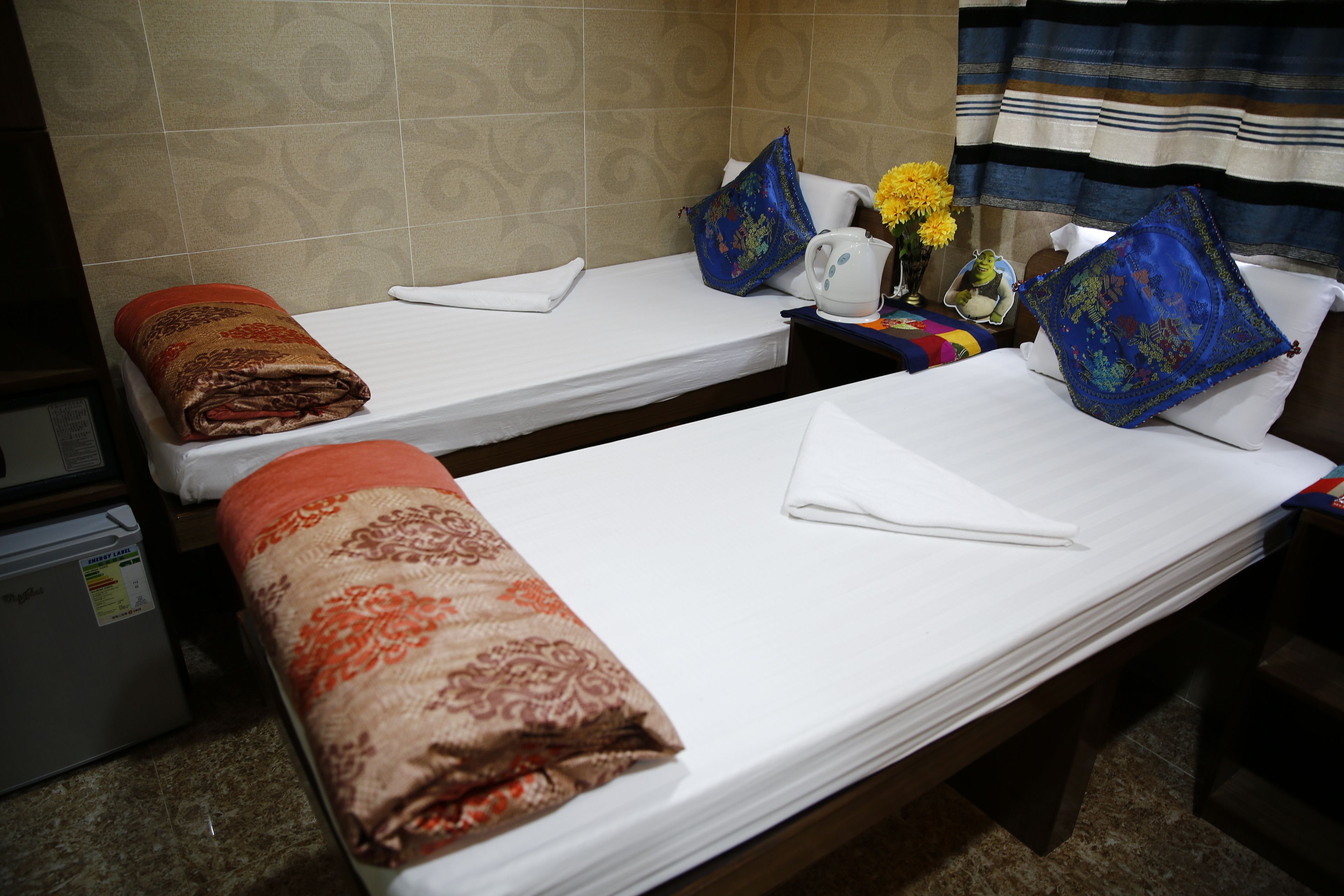 Two single beds with pillows and blankets, a small table with a vase of flowers, and a wall with patterned tiles.
