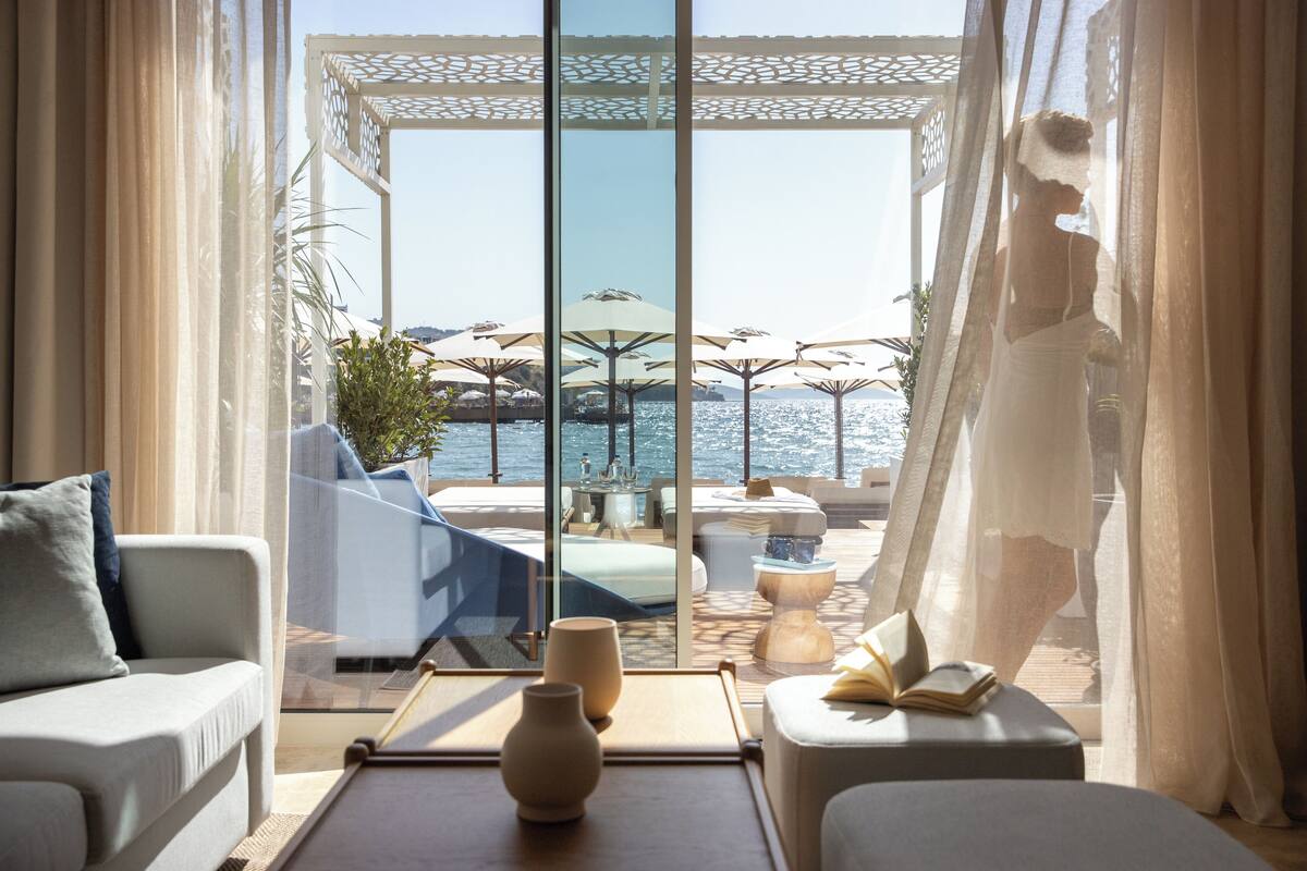 Een moderne hotelkamer met een grote glazen deur die leidt naar een terras met parasols en uitzicht op zee.