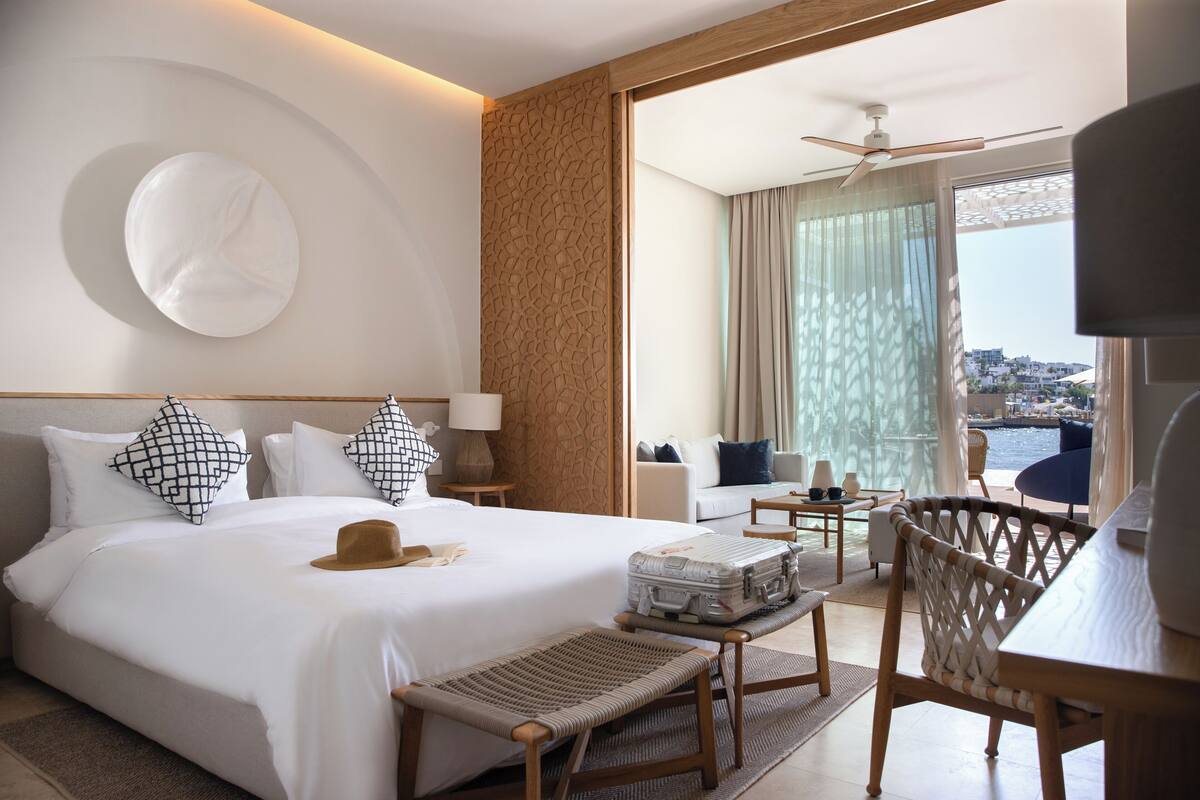 Een hotelkamer met een groot bed, een bureau, een stoel, een koffer en uitzicht op zee.