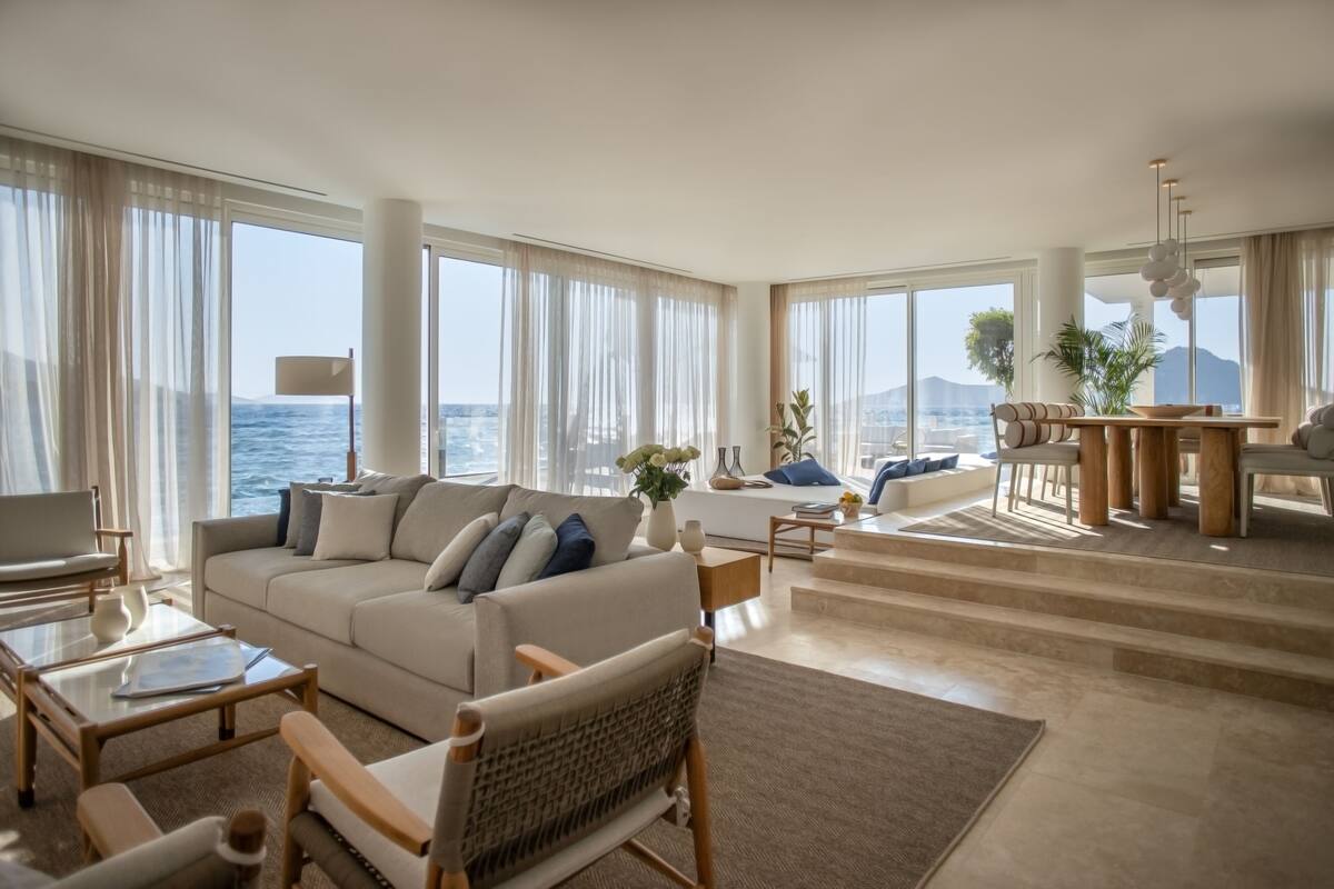 Een ruime woonkamer met een groot raam met uitzicht op zee, een comfortabele bank, een eettafel en een trap naar een hoger gelegen verdieping.