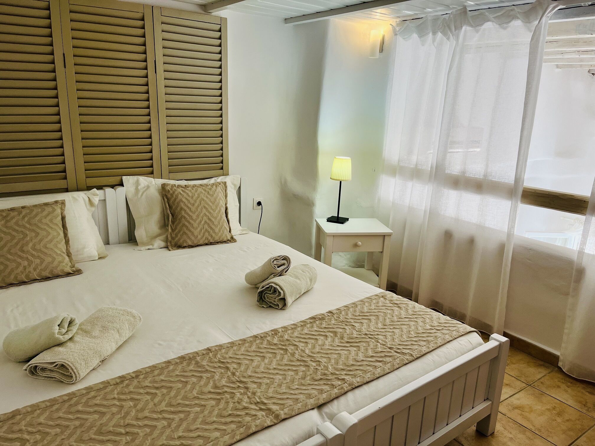 Una cama bien hecha con almohadas y toallas, una mesita de noche con lámpara, y una ventana con cortinas translúcidas.