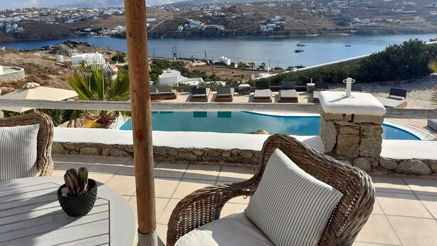 A poolside area with wicker furniture, a thatched umbrella, and a view of a coastal town and sea. - Navy Blue Suites (Mykonos)