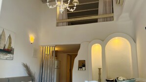 A dining area with a white table and chairs, a chandelier, and a view of the kitchen through an archway. - Navy Blue Suites (Mykonos)