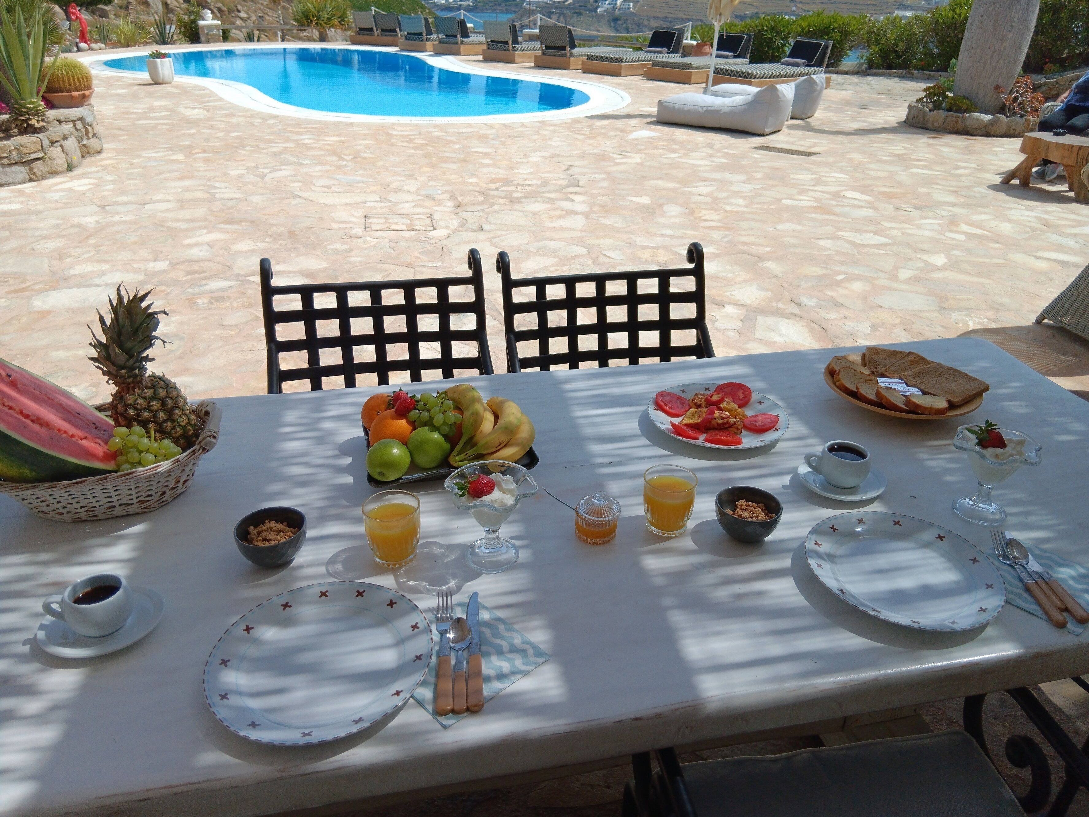 A table set for breakfast with fruits, bread, and beverages, overlooking a swimming pool and outdoor seating area.