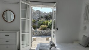 A bedroom with a bed, a dresser, a mirror, and an open door leading to a courtyard with plants and a stone wall. - Navy Blue Suites (Mykonos)